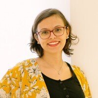 Mulher branca com cabelos escuros na altura do ombro, está usando óculos de grau e vestindo uma blusa preta